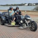 a couple and a driver on a trike with helmets waving with the Mandurah Estuary in the background